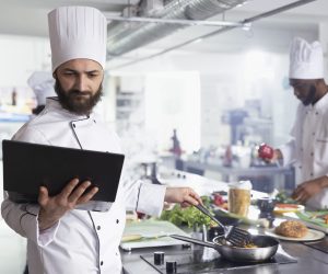 In a restaurant kitchen, male chef in a uniform holds a laptop researching recipes while doing food prep with vegetables and other ingredients. Cooking gourmet dishes on the stove.